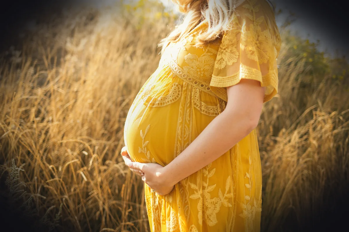 pregnant woman wearing yellow floral dress standing while touching her tummy and facing her right side near brown field durin